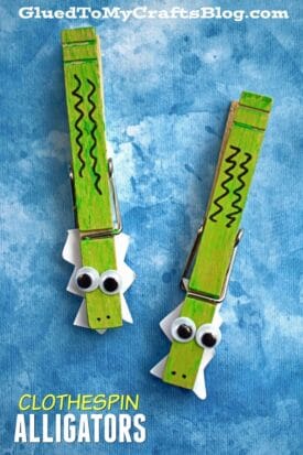 Two wooden clothespins painted green with zigzag lines, googly eyes, and white paper cutouts create a playful clothespin alligator look, displayed on a blue textured background. Text reads CLOTHESPIN ALLIGATORS.