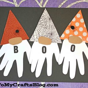 Three handprint Halloween gnomes with white handprint beards, patterned hats, and the letters B, O, O on their bodies spell BOO. Set against a spooky spiderweb background.