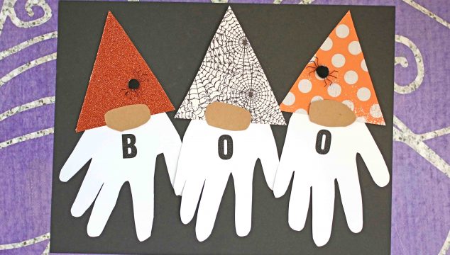 Three handprint Halloween gnomes with white handprint beards, patterned hats, and the letters B, O, O on their bodies spell BOO. Set against a spooky spiderweb background.