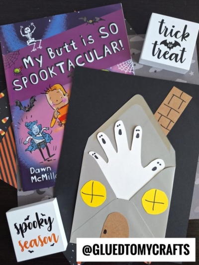 A Halloween-themed flat lay features the book My Butt is SO SPOOKTACULAR!, a handprint ghost in a haunted house craft, and blocks reading trick or treat and spooky season. @GLUEDTOMYCRAFTS tag at the bottom.