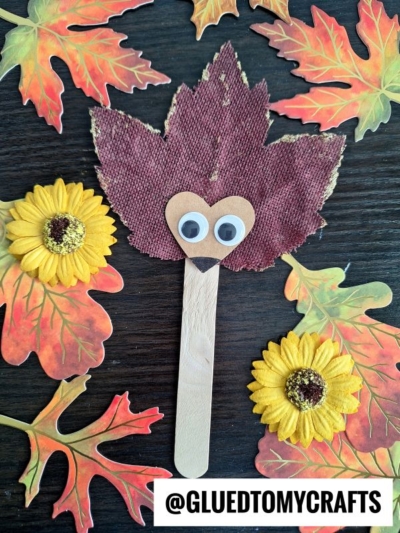 A popsicle stick craft shaped like a leaf hedgehog with googly eyes and a heart-shaped nose, surrounded by artificial autumn leaves and yellow flowers on a dark surface. A label at the bottom reads @GLUEDTOMYCRAFTS.