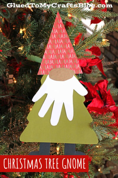 A handmade Christmas tree gnome ornament with a red hat, white beard made from a paper handprint, and green tree-shaped body hangs on a decorated Christmas tree with red flowers. A charming christmas gnome for your festive decor. Text: CHRISTMAS TREE GNOME.