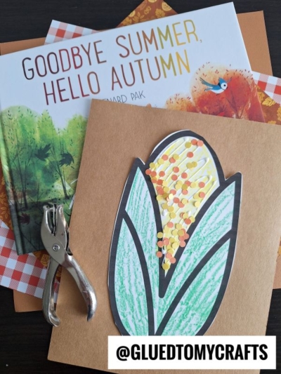 A handmade paper Indian corn craft with colored dots on brown paper sits beside a hole punch, a book titled Goodbye Summer, Hello Autumn, and orange gingham paper. Text reads @GLUEDTOMYCRAFTS.