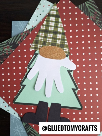A handmade tree gnome craft featuring a green tree-shaped body, plaid hat, white hand-shaped beard, black boots, and a glittery oval nose. Background displays layered patterned papers. Text: @GLUEDTOMYCRAFTS.