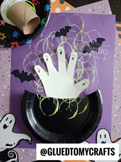 A Halloween craft features a handprint ghost on purple paper, surrounded by bats, curly green lines, and a Paper Plate Witch Cauldron. Cartoon ghosts and a cardboard tube are nearby. Text reads @GLUEDTOMYCRAFTS.