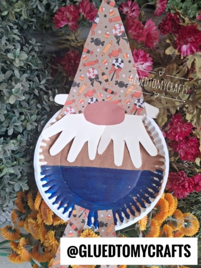 A paper plate fall gnome craft made from a painted paper plate and a patterned paper hat is displayed on flowers and greenery. The hat features festive Halloween designs, adding seasonal charm to the project. The text @GLUEDTOMYCRAFTS appears at the bottom.