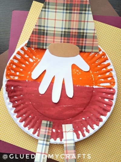 A paper plate fall gnome craft featuring a red and orange painted face, a plaid paper hat and legs, and a white handprint for a beard, all arranged on colorful autumn backgrounds. This charming autumn gnome is perfect for celebrating the season with creativity!.