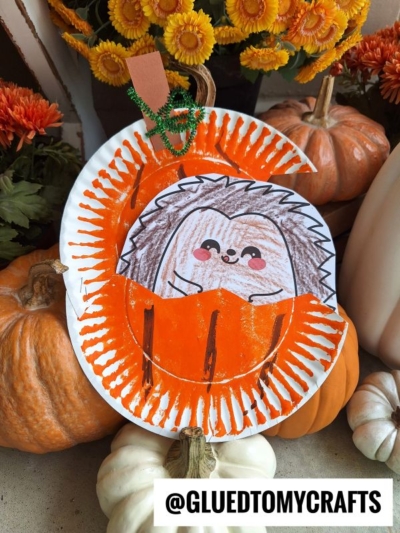 A paper plate pumpkin craft showcases a “hedgehog in pumpkin” design, with an orange painted plate and a cut-out center revealing a cute colored hedgehog. Surrounded by real pumpkins and orange flowers. Text: @GLUEDTOMYCRAFTS.