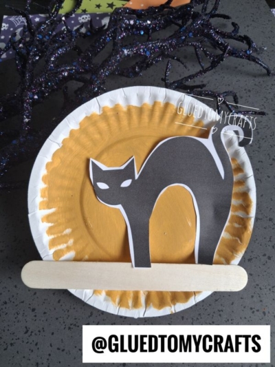 A paper plate painted yellow with a cutout black cat and a craft stick, set on a gray surface—perfect for cat in the night craft or other halloween night crafts. GLUEDTOMYCRAFTS is written on the plate and at the bottom of the image.