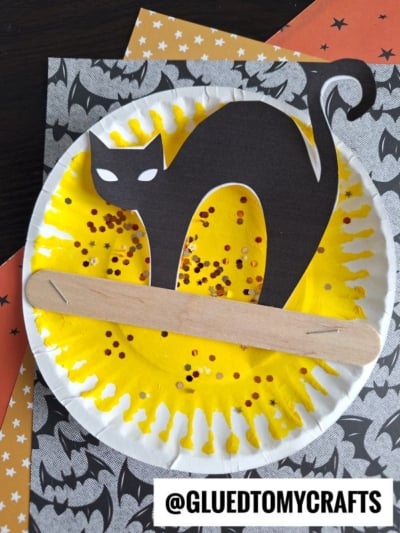 A paper plate painted yellow with orange glitter, featuring a black cat cutout and a craft stick across it, is placed on patterned paper—perfect for cat in the night craft or other fun Halloween night crafts. The handle @GLUEDTOMYCRAFTS appears at the bottom.