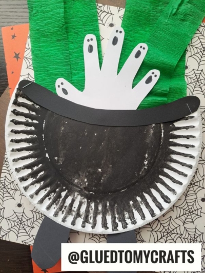 A Halloween craft featuring a Paper Plate Witch Cauldron, green crepe paper strips, and white handprints with drawn-on black fingernails, creating a spooky witch effect. @GLUEDTOMYCRAFTS is written at the bottom.