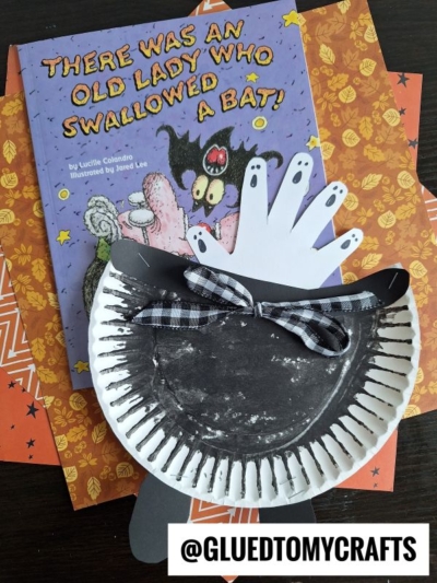 A children’s book titled “There Was an Old Lady Who Swallowed a Bat!” sits on orange Halloween-patterned paper. In front is a black paper plate bat craft, joined by a playful Paper Plate Witch Cauldron for spooky, festive fun.