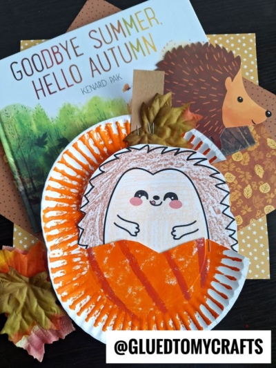 A children’s book titled “Goodbye Summer, Hello Autumn” lies next to a paper plate craft of a smiling hedgehog using orange paint and brown coloring, with autumn leaves and a cute card showing a hedgehog in pumpkin nearby.