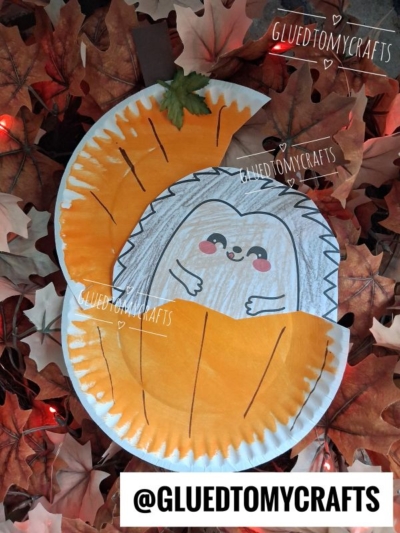 Paper plate craft of a smiling hedgehog in pumpkin, peeking out of an orange pumpkin, surrounded by artificial autumn leaves and small lights. @GLUEDTOMYCRAFTS is written in white text in several places.