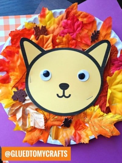 A paper plate decorated with colorful artificial fall leaves forms a playful leaf lion mane around a cute cat face made of yellow and black paper with googly eyes. The image has an orange @GLUEDTOMYCRAFTS label in the corner.