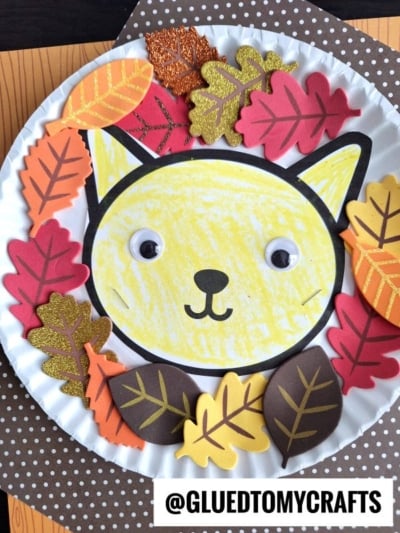 A paper plate craft featuring a cat face colored yellow with googly eyes, surrounded by colorful paper and glittery leaf lion mane on a brown polka-dot background. @GLUEDTOMYCRAFTS is printed at the bottom.