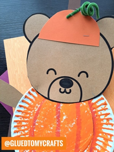 A cute paper plate teddy bear craft made from paper and a painted plate, wearing an orange pumpkin hat with a green pipe cleaner stem. This adorable jack-o-lantern teddy bear is perfect for Halloween fun! @GLUEDTOMYCRAFTS tag is visible in the corner.