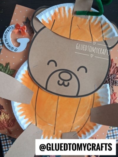 A paper plate teddy bear with an orange pumpkin body made from a painted paper plate, surrounded by fall leaves. The jack-o-lantern teddy bear is smiling, and there’s a GLUEDTOMYCRAFTS watermark and handle at the bottom.