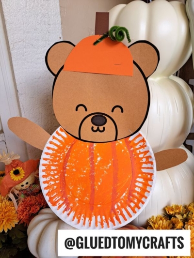A paper plate teddy bear craft with a jack-o-lantern orange pumpkin body, brown arms, and a pumpkin hat, displayed among autumn flowers and white pumpkins. The Instagram handle @GLUEDTOMYCRAFTS appears at the bottom.
