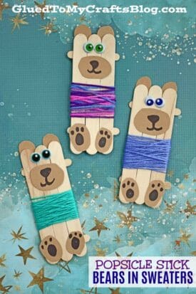 Three popsicle stick bears in sweaters, each with googly eyes, brown paper muzzles and paws, and colorful yarn outfits, sit on a teal background decorated with gold star accents. Text reads: “Popsicle Stick Bears in Sweaters.”.
