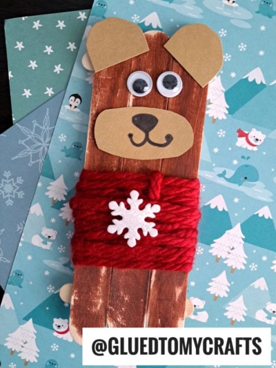 A bear craft made from brown popsicle sticks with googly eyes, a paper nose and ears, and red yarn wrapped like a cozy sweater—perfect popsicle stick bears in sweaters! Displayed on winter-themed paper. Text reads @GLUEDTOMYCRAFTS.