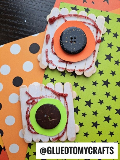 Two popsicle stick eyeball crafts designed as monster eyes with large buttons in the center, glitter glue accents, and colorful paper backgrounds in orange and green Halloween patterns. @GLUEDTOMYCRAFTS is at the bottom.