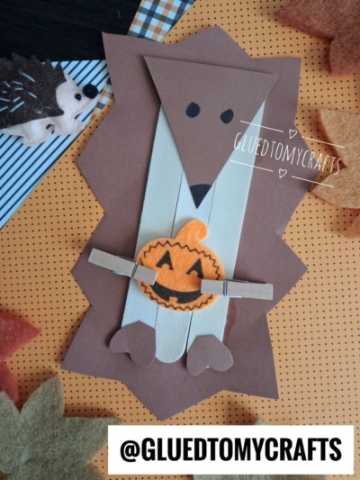 A handmade popsicle stick hedgehog made with felt, holding a small felt pumpkin with clothespins.
