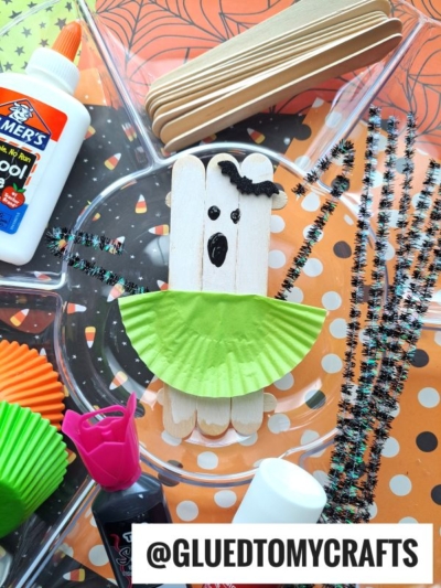 A ghost craft made from popsicle sticks, a green cupcake liner, black pipe cleaners, glue, and markers—these charming popsicle stick ghost ballerinas are displayed on a Halloween-themed tray.