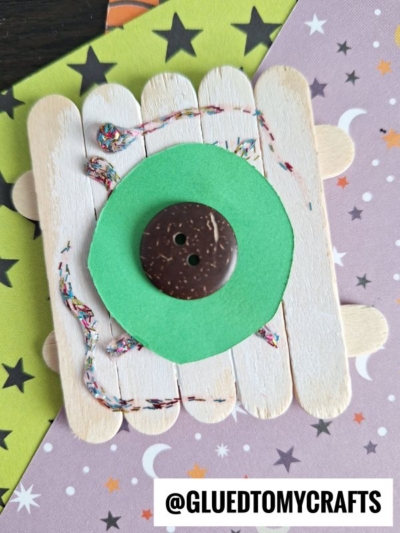 A craft project made of popsicle sticks forms a square base topped with a green paper circle and a brown button, resembling a popsicle stick eyeball. Colorful yarn winds around the sticks atop star- and moon-patterned paper.