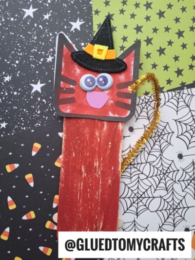 A handmade Halloween popsicle stick cat bookmark shaped like a red cat with googly eyes and a witch hat, decorated with an orange pipe cleaner tail, on a background of spooky patterned papers. Text reads @GLUEDTOMYCRAFTS.