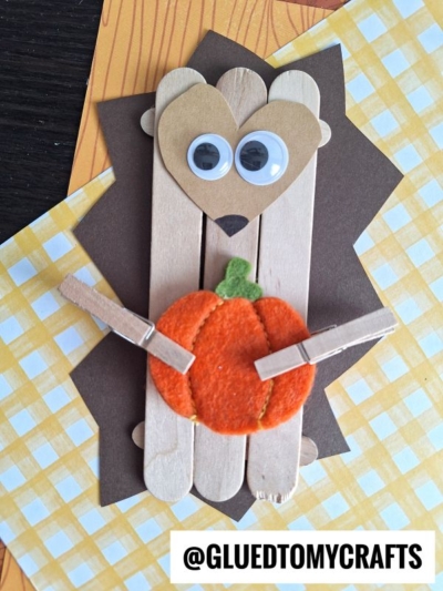 A popsicle stick hedgehog craft featuring a heart-shaped face, googly eyes, and a small felt pumpkin held in its arms. The background is decorated with layered yellow and brown patterned paper for a cozy touch.