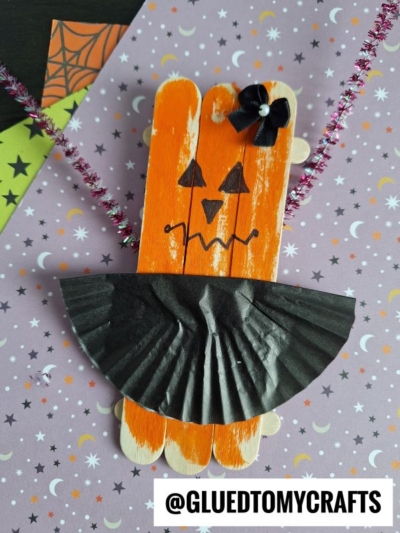 Create adorable Popsicle Stick Jack-O-Lantern Ballerinas by painting popsicle sticks orange, adding a black paper cupcake liner cape, pipe cleaner arms, and a bow. Draw cute faces and display them on festive paper for Halloween flair!.