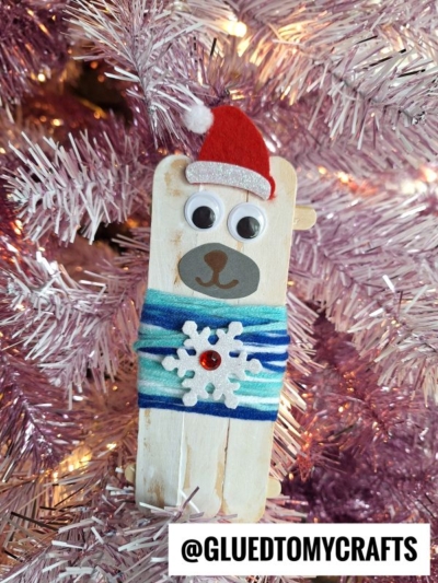 A popsicle stick bear ornament in a cozy sweater, with googly eyes, a red Santa hat, blue yarn, and a white snowflake detail, hangs on a pink Christmas tree. @GLUEDTOMYCRAFTS is written at the bottom.