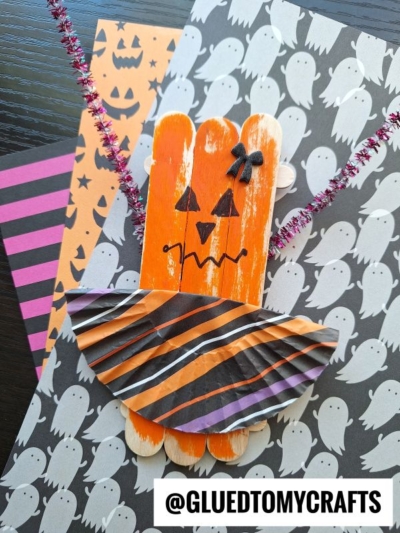 A Halloween craft featuring an orange popsicle stick witch with pipe cleaner arms, a black bow, and a striped cupcake liner hat—similar in spirit to Popsicle Stick Jack-O-Lantern Ballerinas—set on ghost and pumpkin themed papers. @GLUEDTOMYCRAFTS.