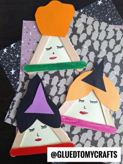 Three popsicle stick Sanderson Sisters witch faces, crafted with colored paper and markers, sit on Halloween-themed ghost and star paper. Each has a unique hat and hair color. @GLUEDTOMYCRAFTS is written at the bottom.