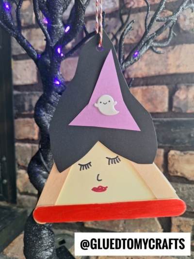A Halloween craft of a witch’s face made from popsicle sticks, inspired by the popsicle stick Sanderson sisters, featuring black hair, a purple hat with a cute white ghost, closed eyes, and red lips. A spooky tree and brick wall are in the background.