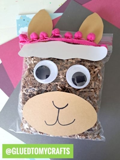 A sealed plastic bag filled with brown crinkle paper creates a charming crinkle paper llama, complete with googly eyes, a paper snout with a drawn mouth, and pink pom-poms. @GLUEDTOMYCRAFTS is written on the bottom left.