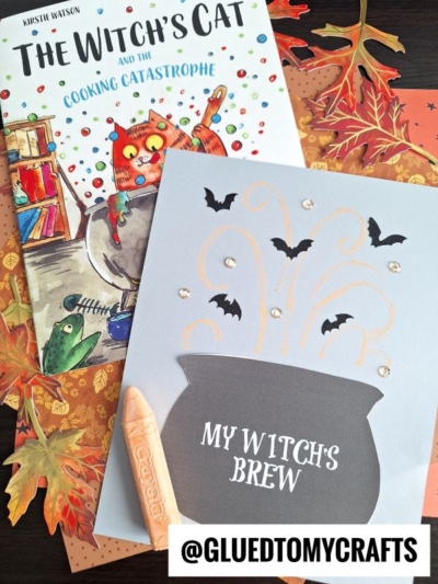 A children's book titled The Witch's Cat and the Cooking Catastrophe sits beside a Halloween-themed craft featuring a chalk art witch cauldron, bats, and swirling lines, all set against a background of autumn leaves.