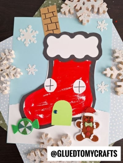 A festive christmas stocking house craft featuring a large red stocking decorated as a cozy home with a green door, black windows, and chimney. Surrounded by snowflakes, candy, and a gingerbread figure on a wintry background.