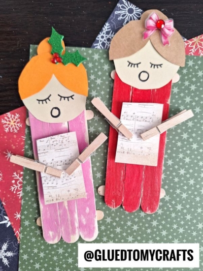 Two handmade popsicle stick Christmas carolers, with paper faces and clothespins holding sheet music, are decorated with bows and holly and lie on festive holiday paper. @GLUEDTOMYCRAFTS is written at the bottom.