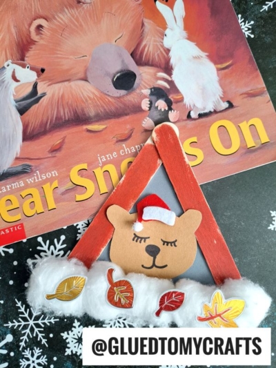 A children’s book titled Bear Snores On lies next to a popsicle stick hibernating bear holiday craft, decorated with cotton, paper leaves, and a red Santa hat. A watermark reads @GLUEDTOMYCRAFTS.