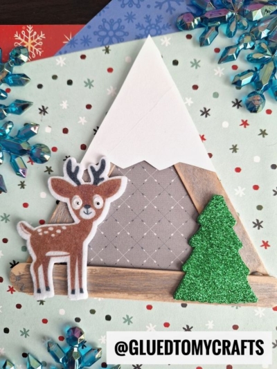 A craft featuring a reindeer, a green glittery tree, and a popsicle stick snowy mountain made from paper and craft sticks, set on a dotted background with blue snowflake decorations and the handle @GLUEDTOMYCRAFTS.