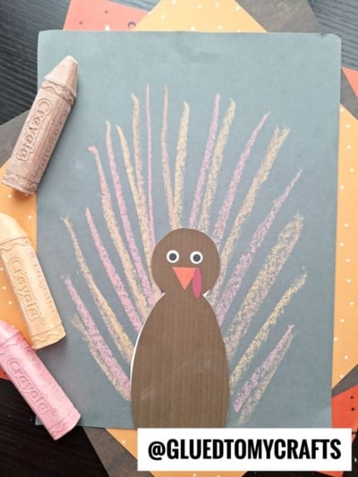 A turkey craft made with paper and crayons shows a brown paper turkey with googly eyes and a red wattle. Its tail feathers are drawn with pink, yellow, and orange crayon lines, adding vibrant color reminiscent of a chalk art turkey. Crayons lie beside the artwork.