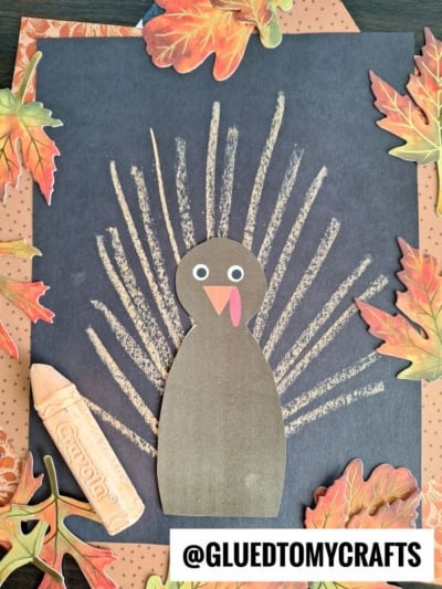 A brown paper turkey with googly eyes is glued to black paper. Yellow crayon lines form feathers behind it, adding a fun chalk art turkey vibe. Decorative autumn leaves and a peach crayon are around the border. Text reads @GLUEDTOMYCRAFTS.