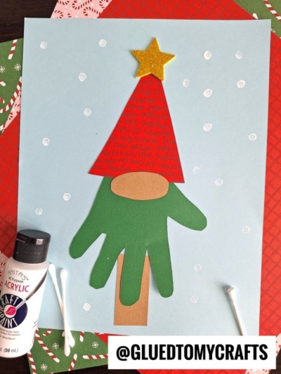 A handprint Christmas tree gnome craft made from a green handprint cutout, brown trunk, red triangle hat with yellow star, and snowy white dots on blue paper. Paint, cotton swabs, and paper are nearby for decorating.