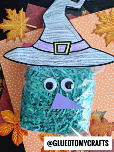 A crinkle paper witch bag filled with green paper strips, decorated as a witch with googly eyes, a purple hat, and a purple paper nose, is placed on orange polka dot paper with fall leaves.