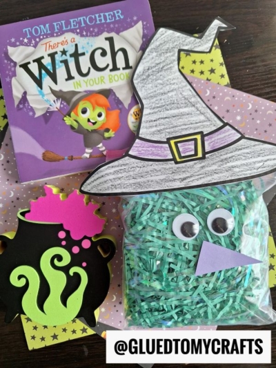 A children’s book titled There’s a Witch in Your Book, a crinkle paper witch hat, a cauldron cutout, and a bag of green shredded paper with googly eyes and a purple triangle nose on a dotted background. @GLUEDTOMYCRAFTS.