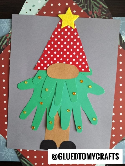 A handprint Christmas tree gnome craft made from colored paper, featuring a red polka dot hat, yellow star, green handprint cutouts for a beard, brown nose and body, and black shoes on gray and red patterned paper.