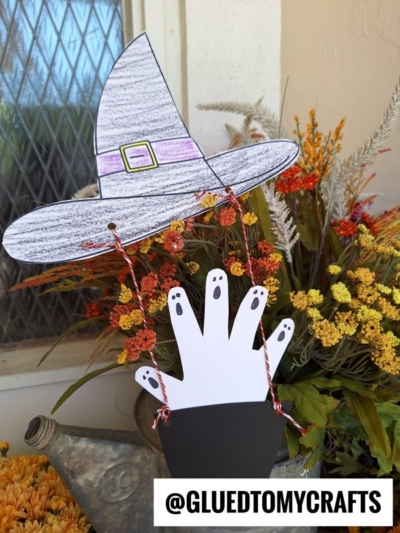 A Halloween craft featuring a cut-out paper witch hat hot air balloon and ghostly white hand, hanging with red string in front of fall flowers. The handle @GLUEDTOMYCRAFTS appears at the bottom right corner.