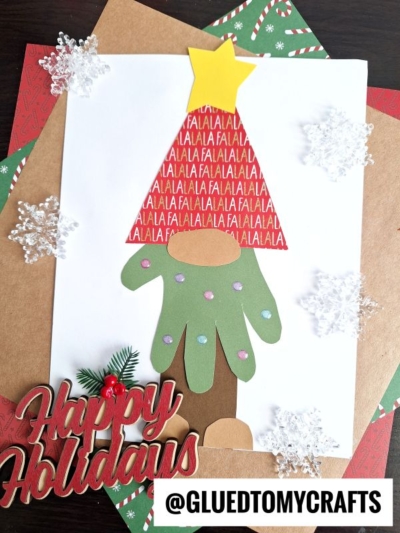 A holiday craft featuring a handprint Christmas tree gnome made from green and brown handprint cutouts, a red triangle with FALALA text, a yellow star on top, paper snowflakes, and Happy Holidays text on festive paper.